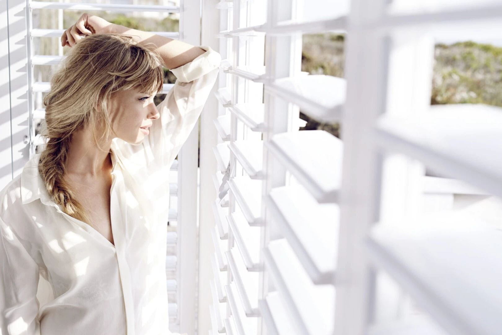 Woman looking through white plantation shutters with natural light filtering in — Seaglass Window Fashions